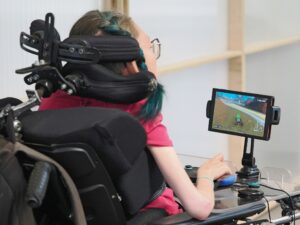 Person im Rollstuhl spielt mit einem Joystick Mario Kart World. Die Kamera schaut ihr perspektiv von hinten über die Schulter