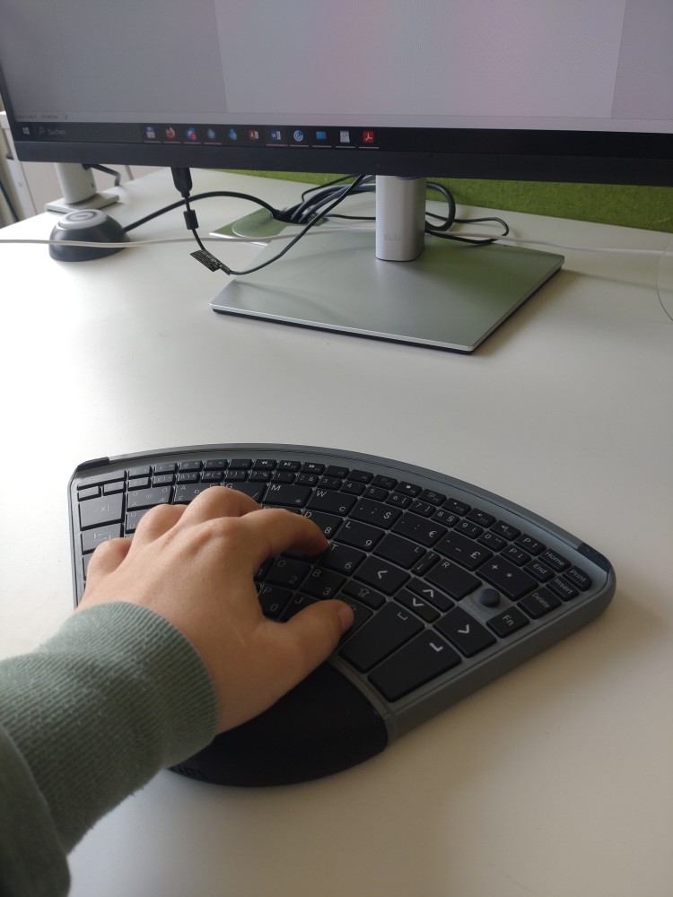 eine mit einer Hand bedienbaren Tastatur in Form eines Kreisausschnitts. Eine Hand ist auf der Tastatur, im Hintergrund ist ein Monitor