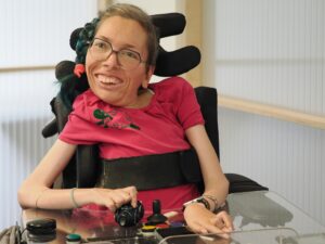Melly lächelt. Sie spielt mit einem Proteus Controler. Weitere adaptive Controler liegen vor ihr auf dem Glastisch.
